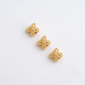 May include: Three gold-colored butterfly-shaped beads. Each bead has a detailed, textured surface with a four-petal design. The beads are arranged diagonally against a white background, showcasing their intricate details.
