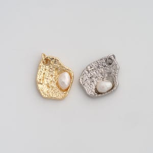 Peut inclure: Deux pendentifs de forme irrégulière, l'un doré et l'autre argenté, chacun orné d'une perle blanche. Les pendentifs ont une surface texturée et une petite boucle en haut pour les suspendre. Le pendentif doré est à gauche, et l'argenté à droite.