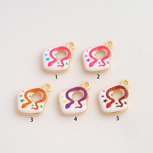 Peut inclure: Cinq breloques en émail blanc avec un contour doré, chacune ornée d'un serpent enroulé et de petites étoiles. La breloque 1 a un serpent rose et des étoiles bleues, la 2 un serpent rose et des étoiles violettes, la 3 un serpent orange et des étoiles vertes, la 4 un serpent violet et des étoiles jaunes, la 5 un serpent rouge et des étoiles noires.