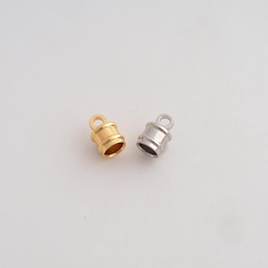 May include: Two cylindrical cord end caps for jewelry making, one finished in a polished gold tone and the other in a polished silver tone. Each cap features a smooth, rounded body with a small circular loop at the top for attachment purposes.