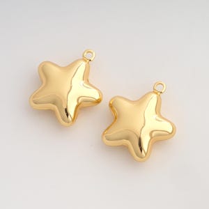 May include: Two gold-colored star-shaped charms with a smooth, reflective metallic finish. Each charm features a small circular loop at the top point, designed for attachment to jewelry or other decorative items, showcasing a simple five-pointed shape.