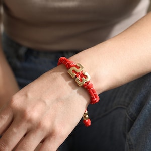 May include: A red cord bracelet featuring a polished gold-tone metal charm in the shape of the number 23. The bracelet is constructed from braided red cord and includes a small gold-tone bead at the end of the adjustable closure mechanism.