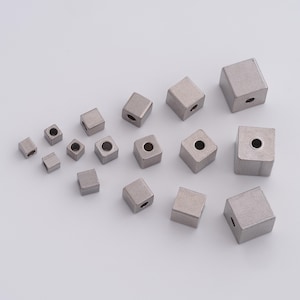 Könnte beinhalten: Eine Sammlung kleiner, quadratischer Metallperlen mit einem Loch in der Mitte. Die Perlen sind in verschiedenen Größen erhältlich und haben eine gebürstete Metalloberfläche. Die Anordnung ist verstreut und zeigt die verschiedenen Größen.