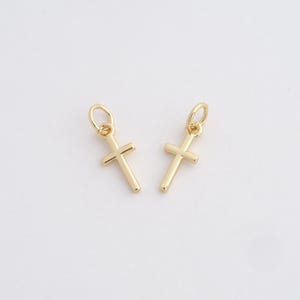 Puede incluir: Dos colgantes con forma de cruz dorada con pequeños bucles en la parte superior para colgar. Las cruces tienen un diseño simple y clásico, con una barra horizontal que cruza una barra vertical. El fondo es blanco liso.