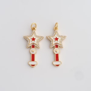 May include: Two gold-colored star-shaped charms with white and red enamel details. Each charm features a star outline encrusted with small clear crystals, a red star center, and a red and white wand-like base. The charms are on a white background.