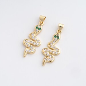 May include: Two gold-coloured snake pendants with clear and green gemstones. The snake bodies are covered in small, clear stones, and the eyes are green. Each pendant has a small loop at the top for attaching to a chain or other jewellery.