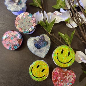 May include: Assortment of colorful phone grips with various designs, including butterflies, smiley faces, and hearts. The grips are made of resin and have a glittery finish.