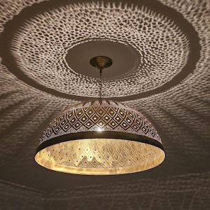 Può includere: Una lampada a sospensione in ottone con un paralume a cupola con intricati intagli geometrici. La luce proietta un'ombra decorativa sul soffitto. La lampada è sospesa a una catena, creando un'atmosfera calda e accogliente.