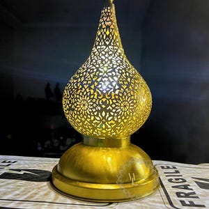 May include: A gold-colored, ornate metal lamp with a teardrop-shaped body and a round base. The lamp's body features intricate cut-out patterns that allow light to shine through. The lamp has a decorative finial at the top.