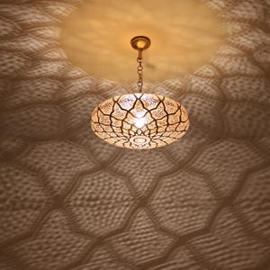 May include: An oval gold pendant light with detailed cut-out designs, suspended from a chain. The light projects a patterned shadow onto the ceiling, creating a warm, decorative atmosphere. The light's intricate design adds a touch of elegance.