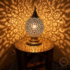 Peut inclure: Une lampe de table sphérique en métal avec un motif floral complexe découpé au laser qui projette des ombres détaillées sur les murs environnants. La lampe présente un sommet pointu, une base circulaire et une lumière interne chaude.
