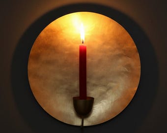 May include: A circular wall-mounted candle holder made of hammered gold-toned metal. A single red taper candle is positioned in the center cup, with a lit flame casting a warm glow against the reflective, textured surface of the circular backplate.