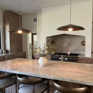 May include: A kitchen island with a light brown countertop and two bar stools. The island is in front of a white kitchen with a brown cabinet and a stainless steel stove. Two large, round, bronze pendant lights hang over the island.
