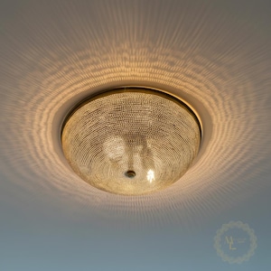 May include: A dome-shaped flush mount ceiling light fixture with a metallic gold finish and a dense, textured surface of small, raised dots. The light emits a warm glow, casting intricate, radiating shadow patterns onto the surrounding ceiling.