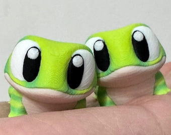 May include: Two small green and white toy gecko figurines with large black eyes. The geckos are posed with their heads tilted up and their mouths slightly open.