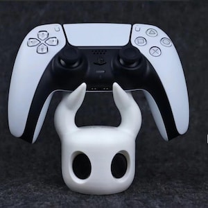 以下が含まれることがあります： 白いPlayStation 5 DualSenseコントローラーが、ホロウナイトというビデオゲームのナイトの形をした白い3Dプリントスタンドの上に置かれています