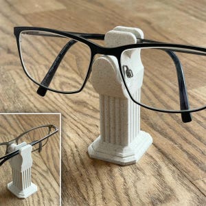 以下が含まれることがあります： 白い斑点のある3Dプリントされた柱型の眼鏡ホルダーに、黒縁の眼鏡が置かれている。