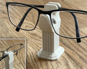 May include: A white, speckled, 3D printed column-shaped glasses holder with a pair of black-framed glasses resting on it.