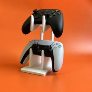 May include: A white plastic stand with two video game controllers. The top controller is black and the bottom controller is white.