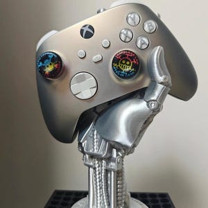 Puede incluir: Mando Xbox plateado con agarres personalizados para el pulgar con diseños de personajes de dibujos animados. El mando está sujeto por un brazo y una base robóticos plateados, creando una pieza de exhibición única. El mando tiene los botones y la cruceta estándar.