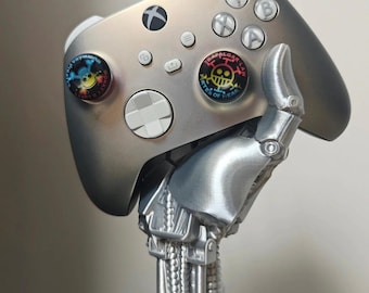 Może przedstawiać: Srebrny kontroler Xbox z niestandardowymi uchwytami na kciuki z postaciami z kreskówek. Kontroler jest trzymany przez srebrne ramię i podstawę robota, tworząc unikalny element ekspozycji. Kontroler ma standardowe przyciski i pad kierunkowy.