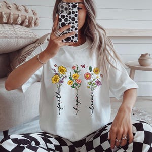 Könnte beinhalten: Weißes T-Shirt mit floralem Design und den Worten "Glaube", "Hoffnung" und "Liebe". Die floralen Arrangements umfassen gelbe Sonnenblumen, rosa und orangefarbene Blumen und lila Blumen. Schmetterlinge sind ebenfalls im Design enthalten.