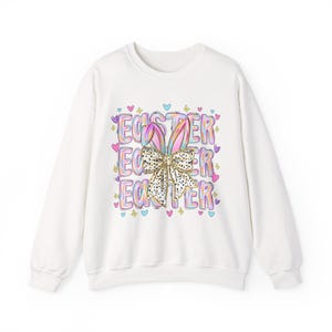 Könnte beinhalten: Weißes Sweatshirt mit einem farbenfrohen Oster-Grafik. Die Grafik zeigt das Wort "EASTER" in einem sich wiederholenden Muster, Hasenohren und eine Schleife. Das Design umfasst Pastellfarben und Herzformen.