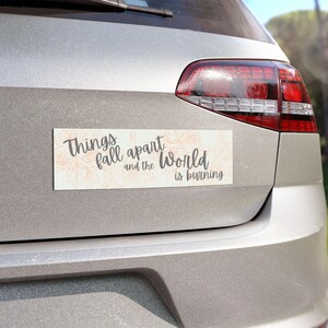 May include: A rectangular bumper sticker with the text "Things fall apart and the World is burning" in gray script. The background features a floral pattern in a light peach color. The sticker is affixed to the back of a silver car.