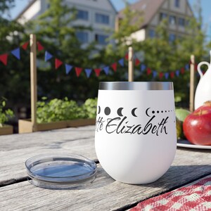 Op de afbeelding: Een witte roestvrijstalen wijnglas met een zwart ontwerp van een halve maan en de tekst "Little Elizabeth". Het glas staat op een houten tafel met een rood-wit geruit tafelkleed.