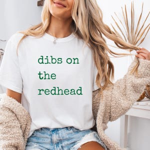 以下が含まれることがあります： 「dibs on the redhead」という緑色の文字が書かれた白いTシャツです。