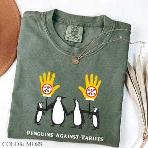May include: A green t-shirt with a graphic of four penguins holding signs that say "Hands Off!" The penguins are wearing yellow gloves. The text "Penguins Against Tariffs" is printed below the penguins. The shirt is labeled "Color: Moss".