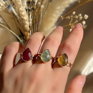 May include: Three gold rings, each with a teardrop-shaped gemstone in red, green, and yellow, are shown on a hand. The rings are set against a backdrop of dried flowers in neutral tones, creating a natural and elegant aesthetic.