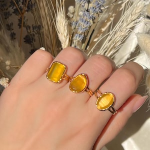 May include: Three gold rings with yellow gemstones are displayed on a hand. The rings feature different gemstone shapes: rectangular, oval, and freeform. The rings are set against a backdrop of dried flowers.