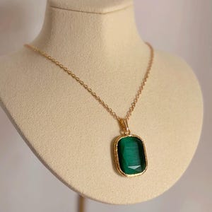Könnte beinhalten: Eine goldfarbene Halskette mit einem rechteckigen, smaragdgrünen Anhänger. Der Anhänger ist in einen goldenen Rahmen gefasst und hängt an einer zarten Goldkette. Die Halskette wird auf einer Büste präsentiert.