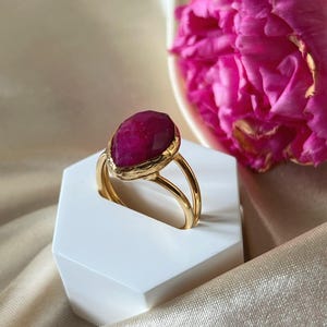 May include: A gold ring featuring a large, faceted, dark pink gemstone. The ring is displayed on a white hexagonal stand.