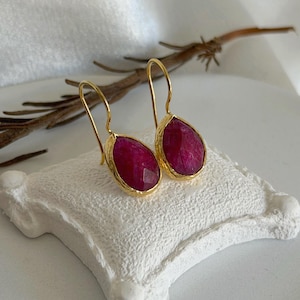 Peut inclure: Une paire de boucles d'oreilles en forme de goutte avec des pierres rose foncé facettées serties dans des montures en métal doré. Les boucles d'oreilles ont des crochets dorés incurvés et sont présentées sur un petit coussin blanc texturé.
