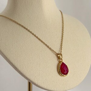 May include: A gold necklace featuring a teardrop-shaped, faceted, red pendant. The pendant is framed in gold and suspended from a delicate gold chain. The necklace is displayed on a cream-coloured bust.