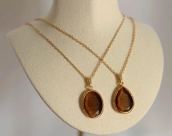 May include: Two gold-colored necklaces with brown gemstone pendants. One pendant is oval, the other is teardrop-shaped. The necklaces are displayed on a cream-colored bust against a soft, neutral background.