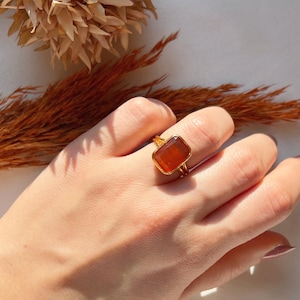 May include: A gold ring featuring a large, rectangular, orange-brown gemstone. The ring is worn on a finger.