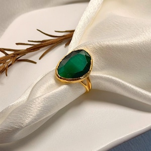 Könnte beinhalten: Ein goldener Ring mit einem großen, smaragdgrünen Edelstein. Der ovale Edelstein ist in einer goldenen Lünette gefasst. Der Ring wird auf einem weißen, seidigen Stoff präsentiert, mit einem Zweig getrockneter Blumen im Hintergrund. Der Ring ist ein Schmuckstück.