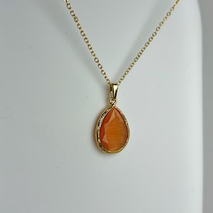 Puede incluir: Un collar dorado con un colgante naranja en forma de lágrima. El colgante tiene una superficie texturizada y está enmarcado por un bisel dorado. La delicada cadena descansa sobre una superficie blanca. El colgante mide unos 2 cm de largo.
