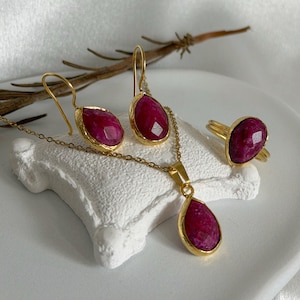 Puede incluir: Un conjunto de joyería que consta de un collar dorado con un colgante de piedra preciosa magenta facetada en forma de lágrima, un par de pendientes a juego en forma de lágrima y un anillo dorado con una piedra magenta facetada en forma de lágrima.