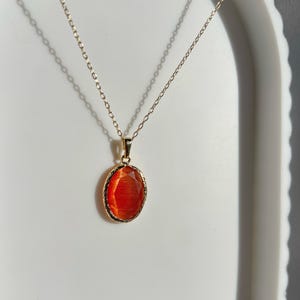 以下が含まれることがあります： 楕円形、オレンジがかった赤色の宝石が付いたゴールドカラーのネックレス。 宝石はゴールドカラーのベゼルにセットされています。 繊細なチェーンが白い表面に掛かっています。 ペンダントの長さは約2cmです。