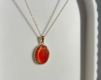 May include: A gold-coloured necklace with an oval, orange-red gemstone pendant. The gemstone is set in a gold-coloured bezel. The delicate chain hangs on a white surface. The pendant is approximately 2 cm in length.