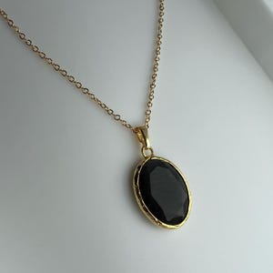 May include: A gold-coloured necklace featuring a black oval pendant. The pendant has a faceted surface and is set in a gold-coloured bezel. The chain is delicate and also gold-coloured. The necklace is shown against a white background.