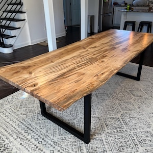 May include: A large wooden dining table with a live edge top and black metal legs. The table is made of wood with a natural grain pattern and is sitting on a patterned rug.