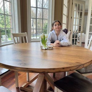 May include: A large round wooden dining table with a natural grain finish and a sturdy cross-base support. The table features a clear glass vase filled with white tulips and a small circular coaster placed in the centre of the tabletop surface.
