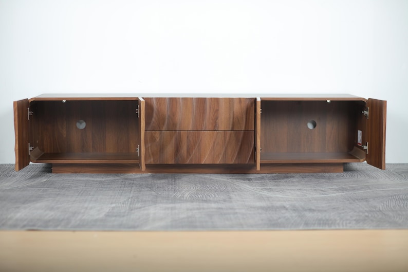 Może przedstawiać: Konsola multimedialna w stylu mid-century modern wykonana z ciemnego drewna. Konsola ma dwoje drzwi po obu stronach, otwieranych, aby odsłonić wnętrze, oraz dwie szuflady pośrodku. Drzwi są otwarte, odsłaniając wnętrze z otworem do zarządzania kablami.