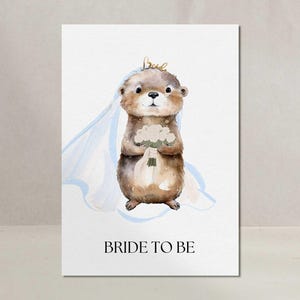 Könnte beinhalten: Eine Aquarellillustration eines braunen Otters, der einen weißen Brautschleier und eine goldene Krone trägt und einen Strauß weißer Blumen hält. Der Text BRIDE TO BE ist in schwarzen Großbuchstaben am unteren Rand der weißen Karte gedruckt.