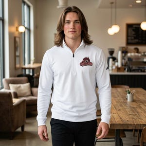 May include: White long-sleeve athletic shirt with a quarter-zip collar. The shirt features a logo on the chest and the Adidas logo on the sleeve. The person is wearing black pants. The background is a cafe.